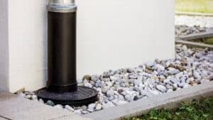 A black downspout extends from a building and drains onto a circular grate, surrounded by white and gray gravel stones and a concrete border.