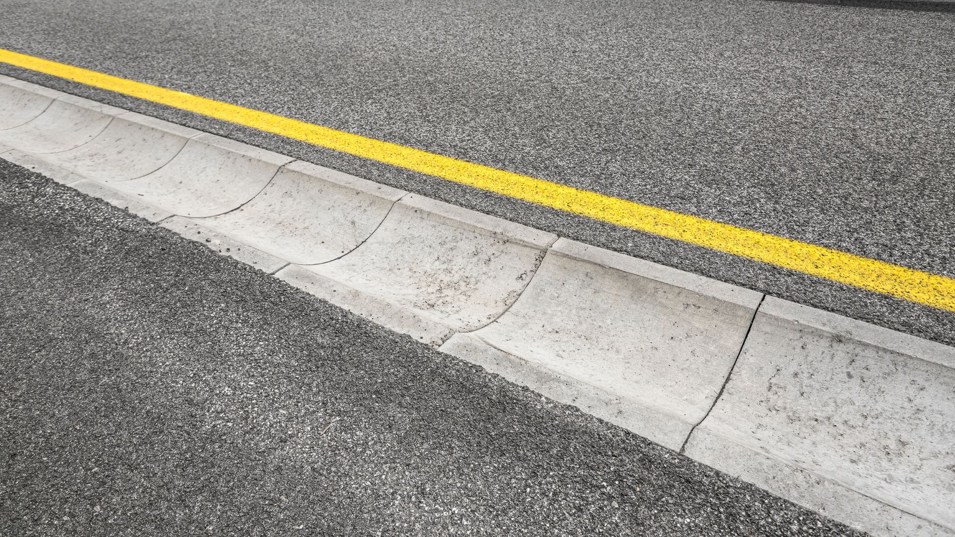 A close-up view of a concrete curb next to an asphalt road with a solid yellow line painted parallel to the curb.