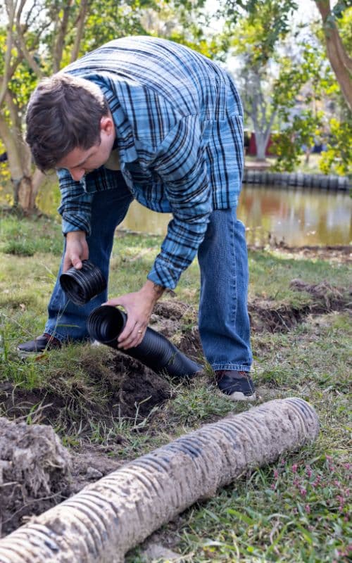 A person outdoors connects two black drainage pipes on a grassy area near a dug trench, with trees and a body of water in the background.