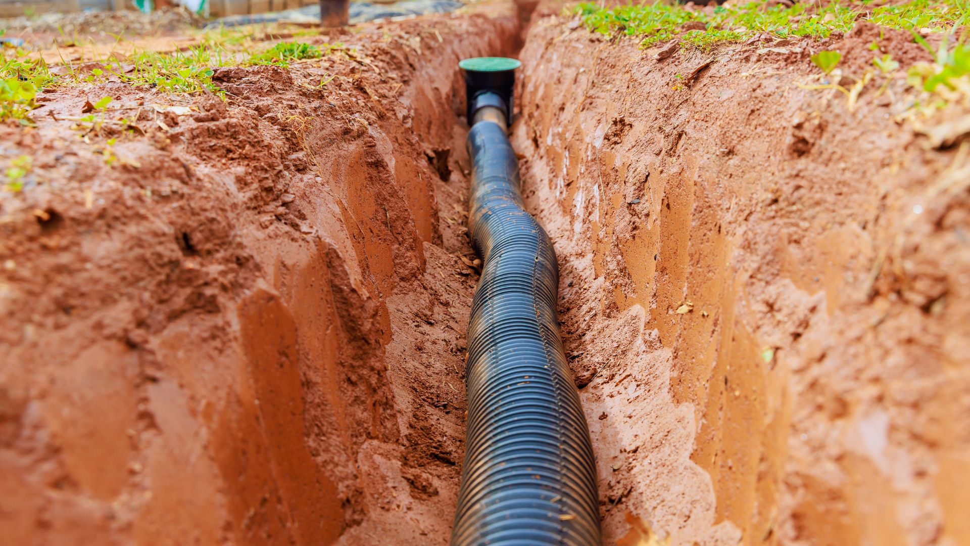 A black corrugated drainage pipe is installed in a narrow trench dug into brown soil, with grass visible along the edges.