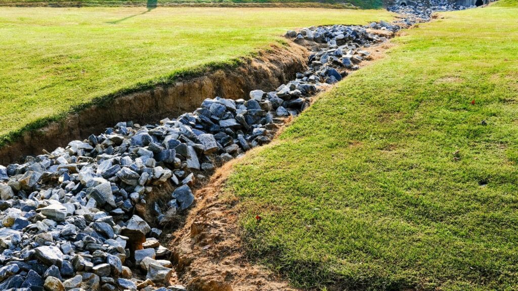 A drainage ditch lined with large rocks runs through a grassy area, with green grass on both sides and a culvert visible in the background.