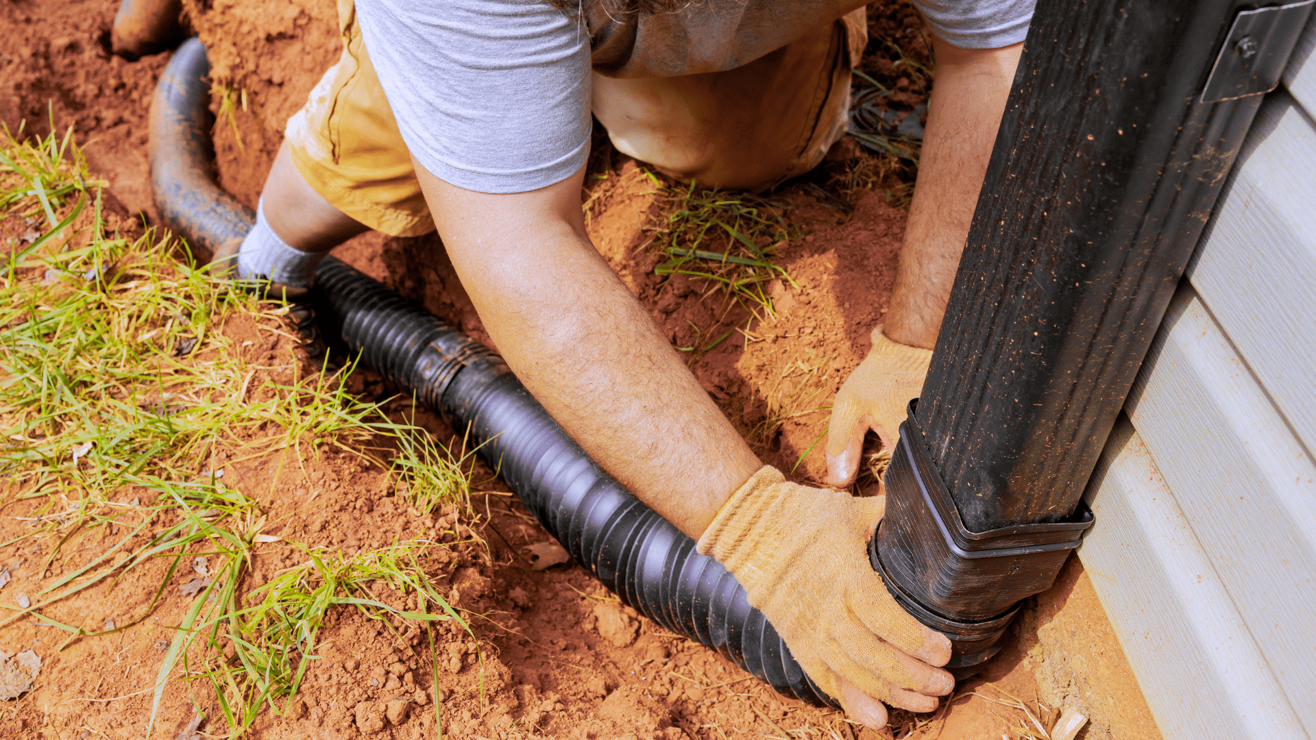 A person wearing gloves connects a black corrugated drainage pipe to a downspout near the base of a building, working on soil.