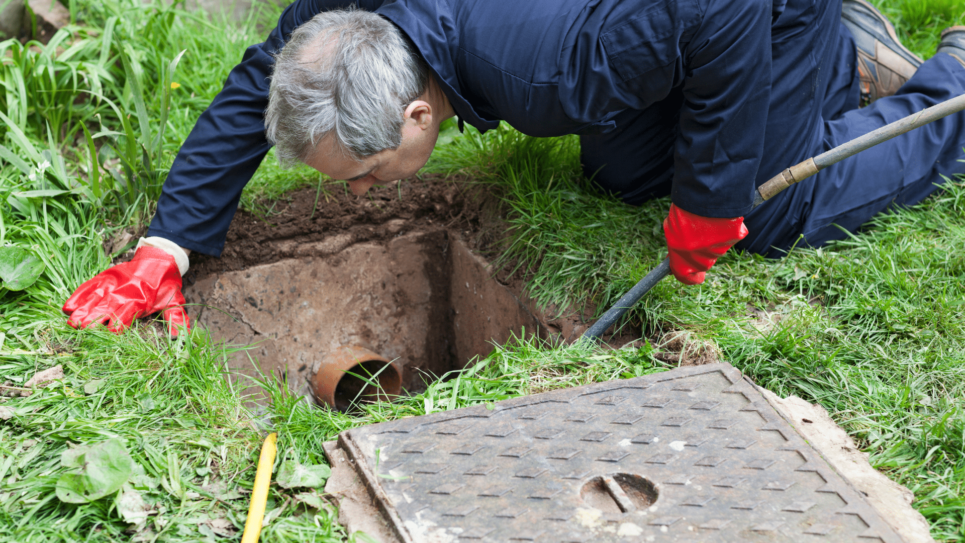 A person wearing red gloves and blue overalls kneels on grass, inspecting an open manhole with a pipe visible inside. A metal cover lies nearby.