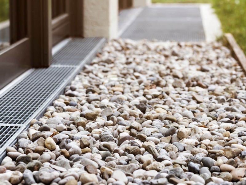 Close-up view of a gravel bed with a metal drainage grate next to the exterior wall of a building. Grass is visible at the edge of the gravel.