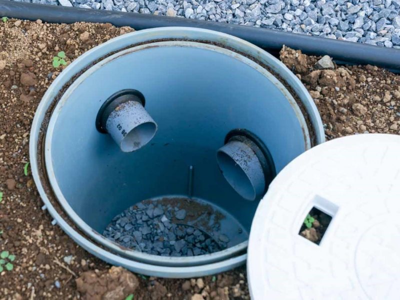 Open inspection chamber with two inlet pipes and a partially removed white cover, surrounded by soil and gravel.