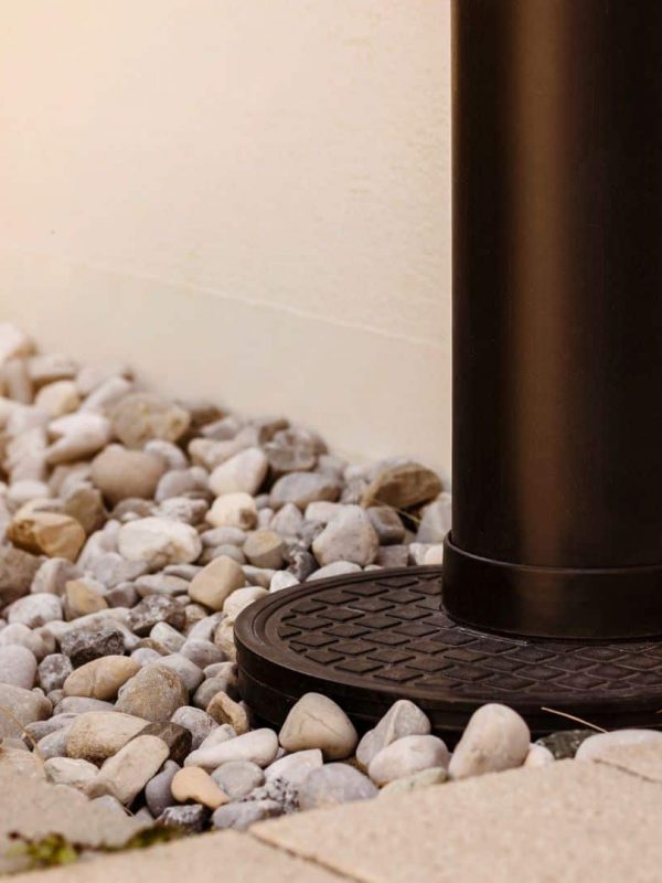 A black drainage pipe extends down a white wall, ending on a circular grate surrounded by white pebbles and paving stones.