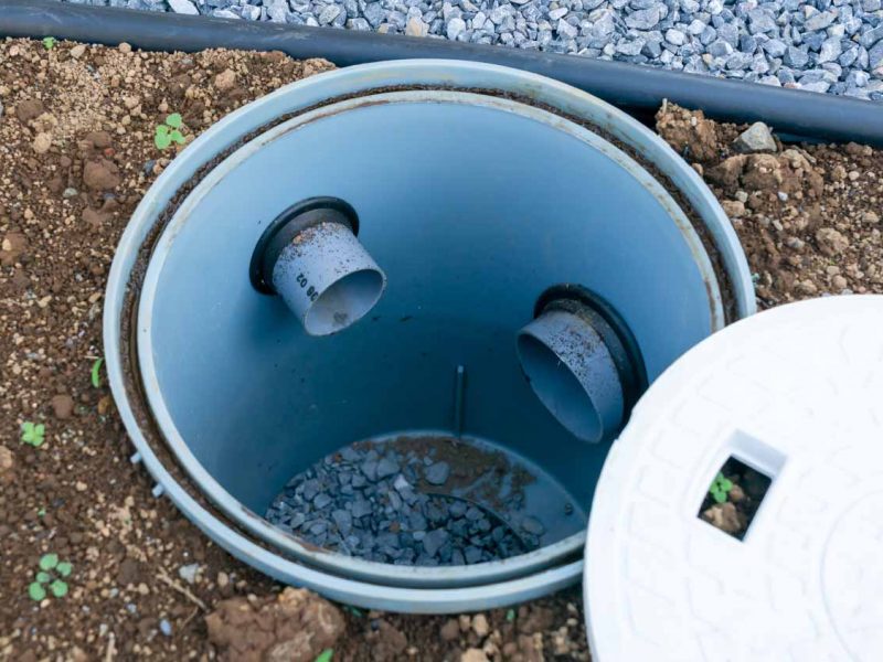 A plastic inspection chamber with two inlet pipes is installed in the ground; the cover is placed nearby on the soil.